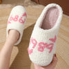 A pair of white plush indoor slippers with pink embroidery are being held against a light wooden background.