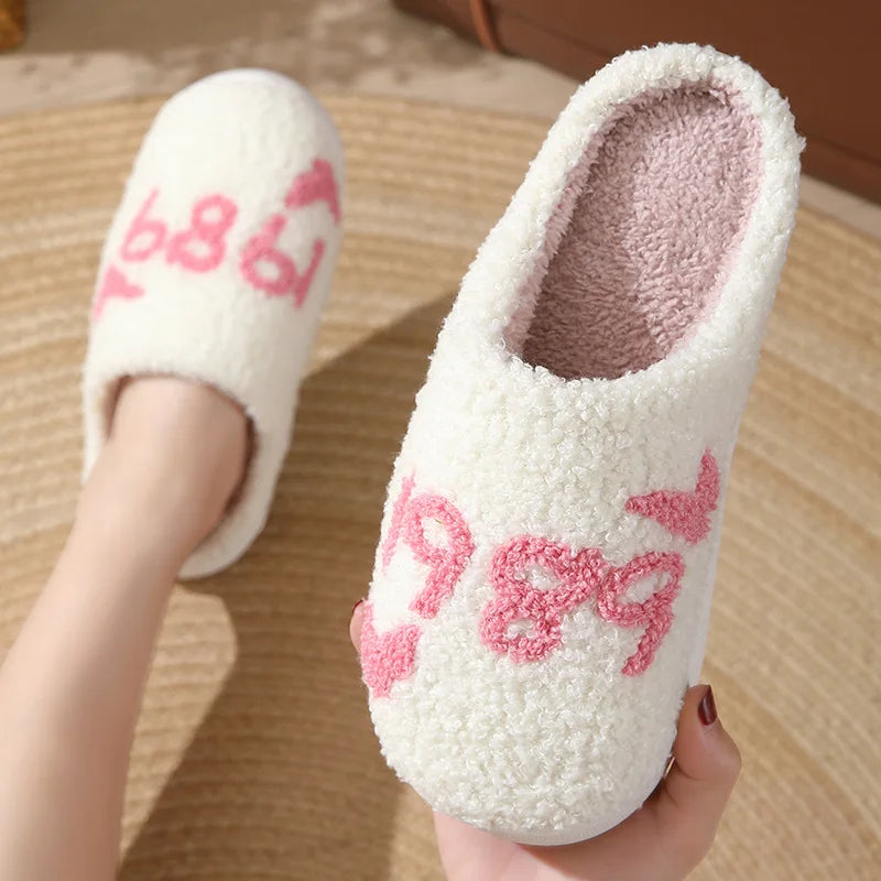 A pair of white plush indoor slippers with pink embroidery are being held against a light wooden background.