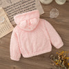 A pink knitted plush hooded winter jacket lying on a wooden surface next to a piece of paper.
