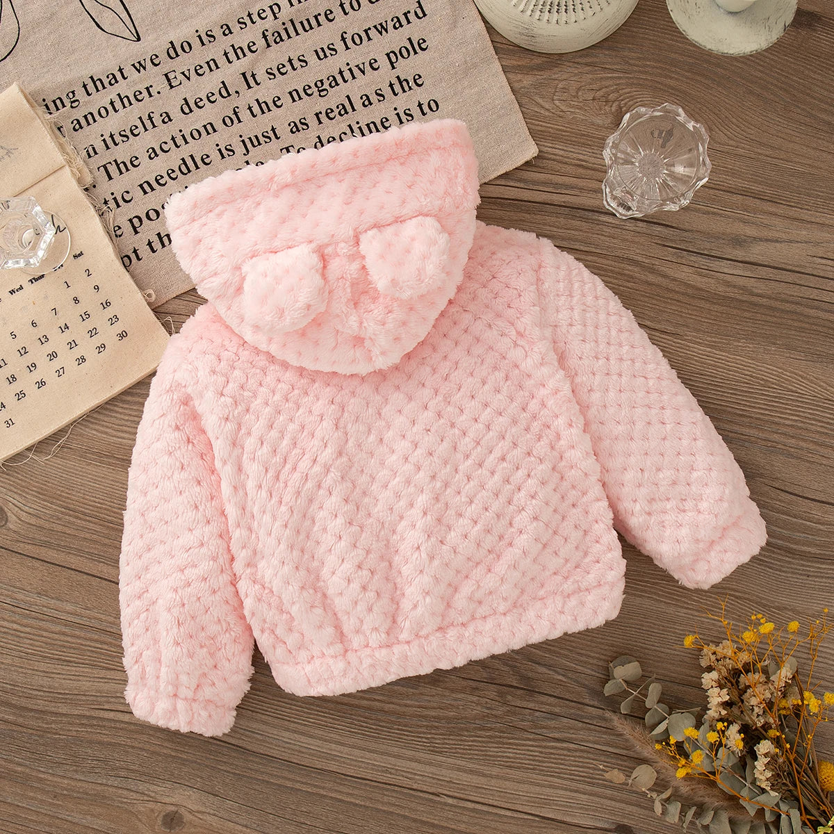 A pink knitted plush hooded winter jacket lying on a wooden surface next to a piece of paper.