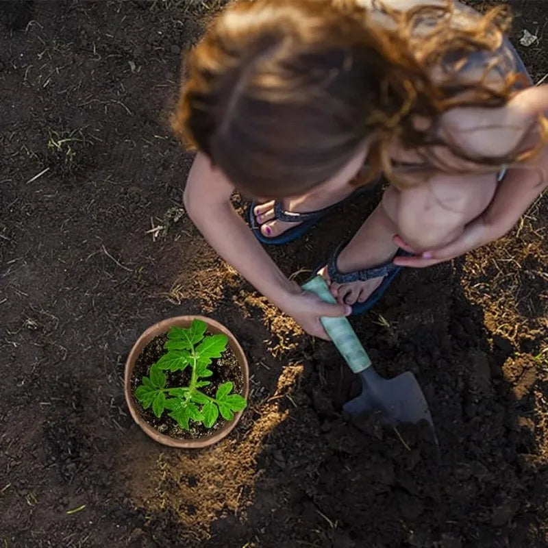 A young person is using a small shovel to dig in dark soil next to a biodegradable peat pot containing green seedlings.