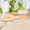 A light wooden baby brush and comb set with an engraved name resting on a beige cloth.