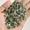 A hand holds a pile of natural tumbled stone chips in various shades of green, brown, and clear quartz.