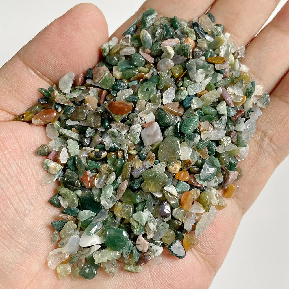 A hand holds a pile of natural tumbled stone chips in various shades of green, brown, and clear quartz.