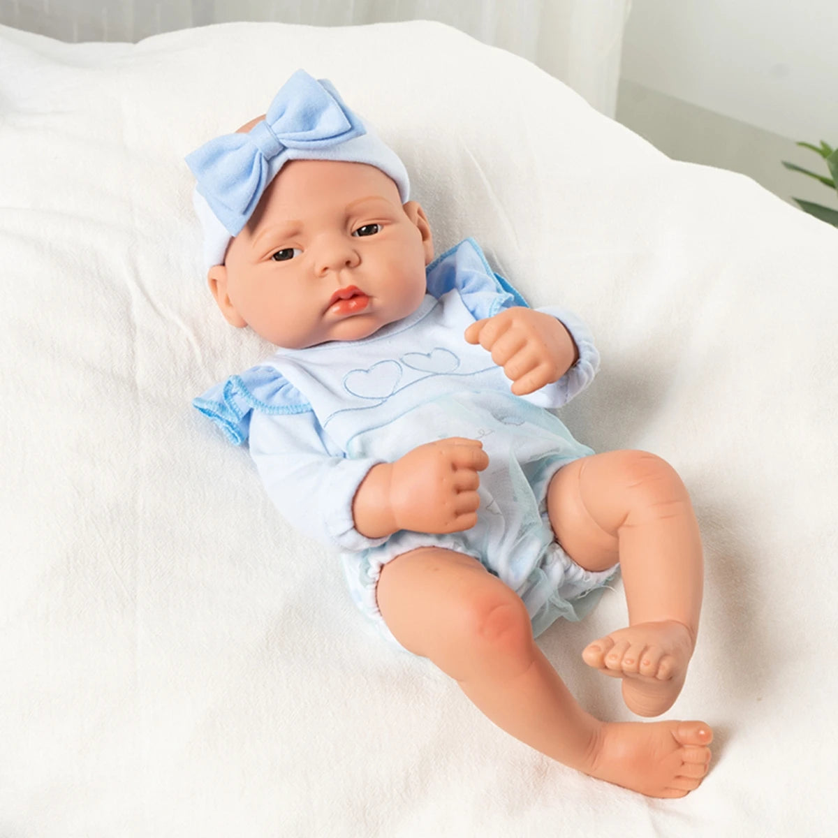 A lifelike vinyl reborn doll with a blue bow headband and matching outfit lying on white fabric.