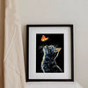 A framed black and white illustration of a cat with glowing yellow eyes and a butterfly on a black background.