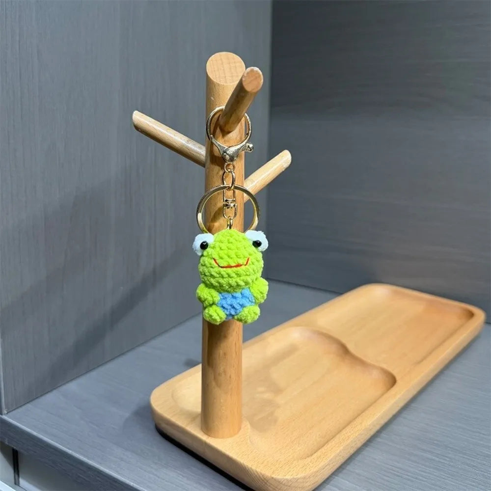 A handmade crochet frog keychain with blue and green yarn, attached to a wooden stick and ring.