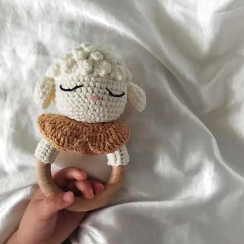 A handmade white knitted baby rattle and teether ring is held in a person's hand against a white fabric background.