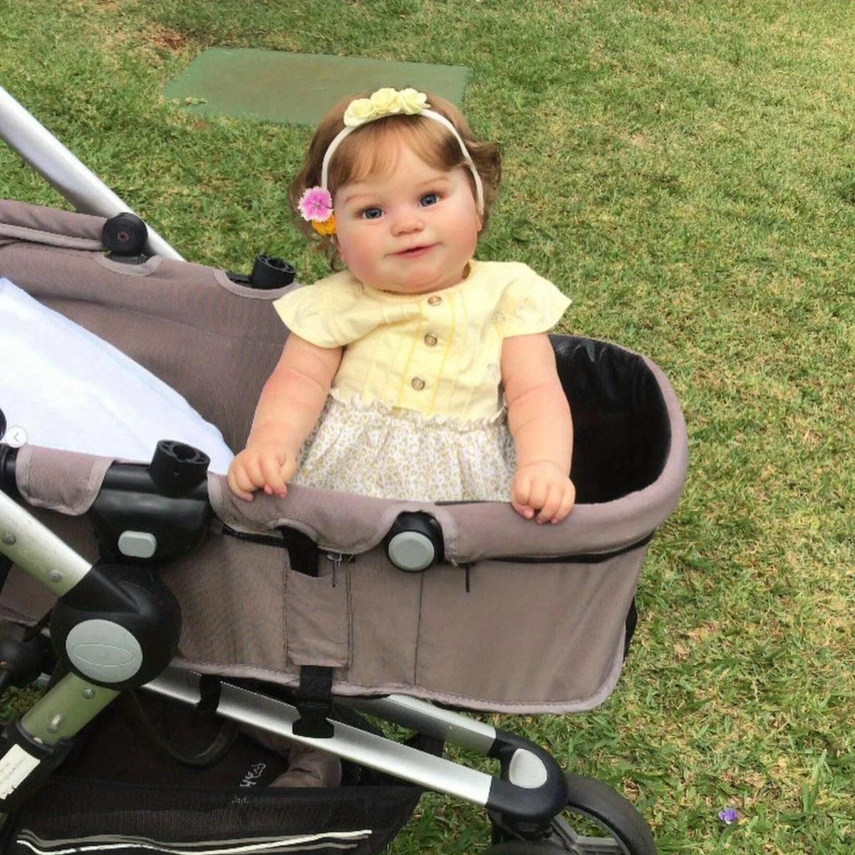 A baby doll with blonde hair and a yellow top is seated in a stroller on a grassy field.