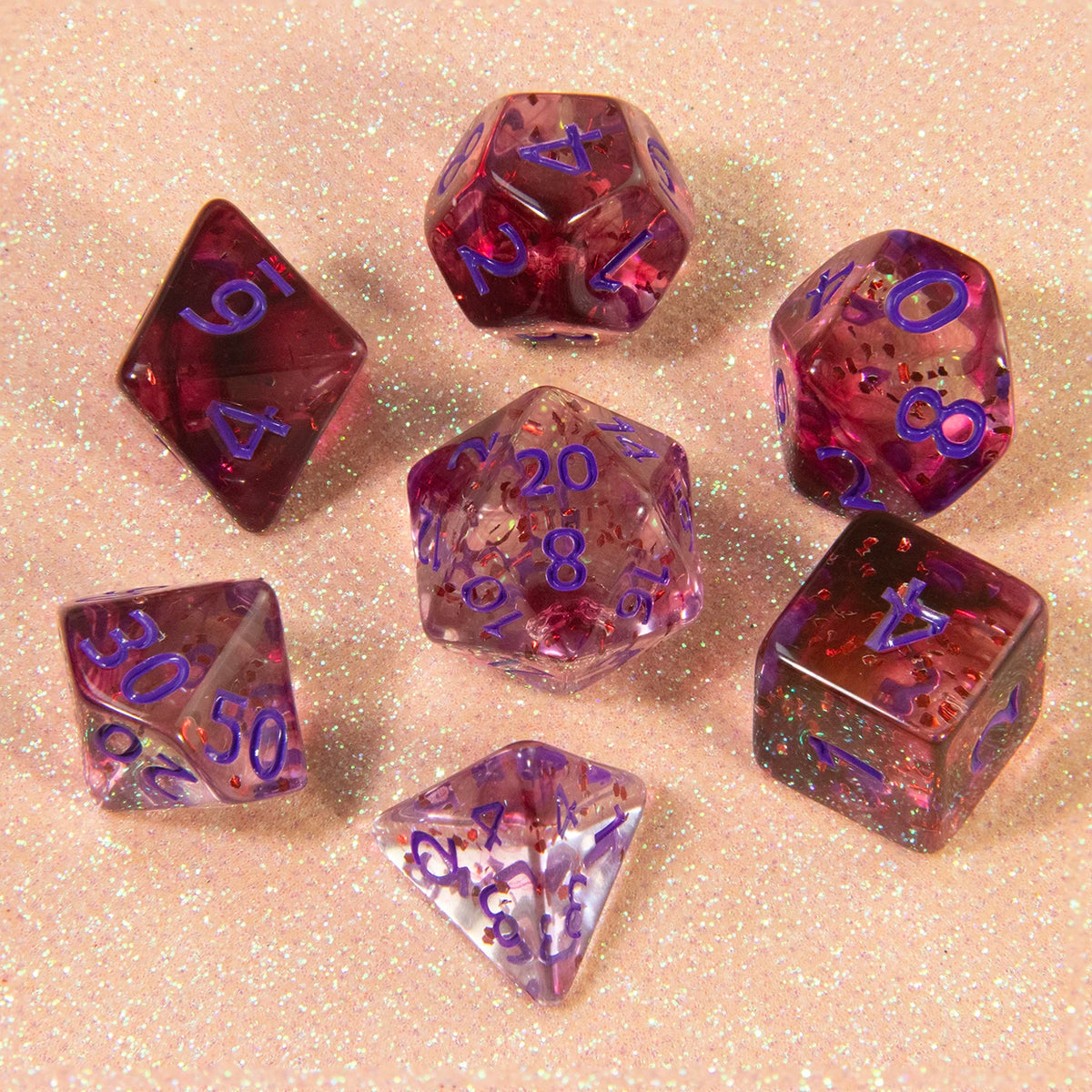 A collection of various colored glitter polyhedral dice including D4, D6, D8, D10, D12, and D20 resting on a pink surface.