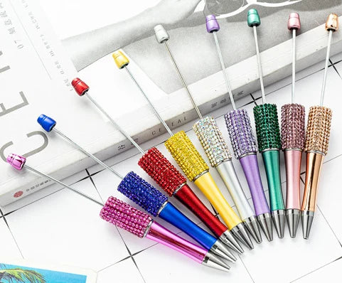 A collection of thirteen colorful beaded ballpoint pens arranged diagonally on a white surface.