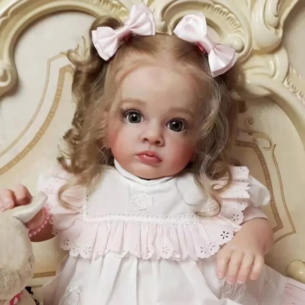 A lifelike reborn baby doll with pink hair and a white dress wearing pink bows.