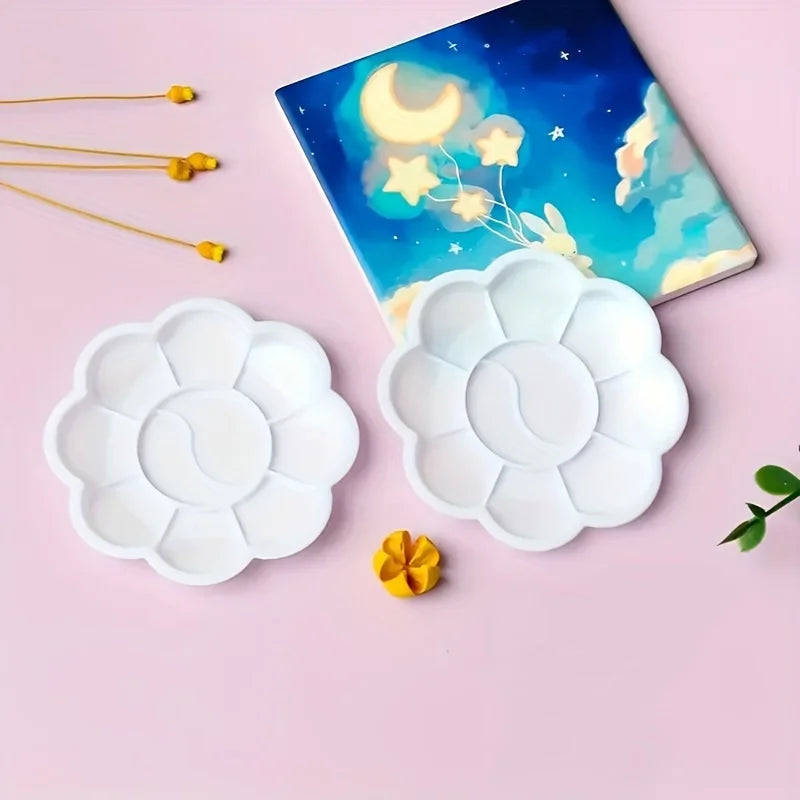 A set of ten flower-shaped white disposable paint palettes rests on a pink surface next to a blue celestial notebook.