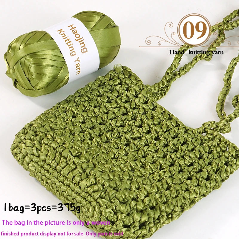 A hand-knitted moss stitch bag made from green satin ribbon yarn with a spool visible.