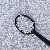 A black spoon holds clear glass crystal rhinestones scattered on a white background.