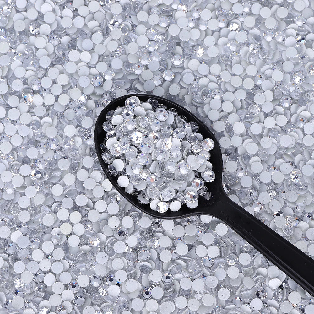 A black spoon holds clear glass crystal rhinestones scattered on a white background.