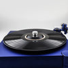 A black aluminum alloy record stabilizer sits on a blue surface next to a turntable.