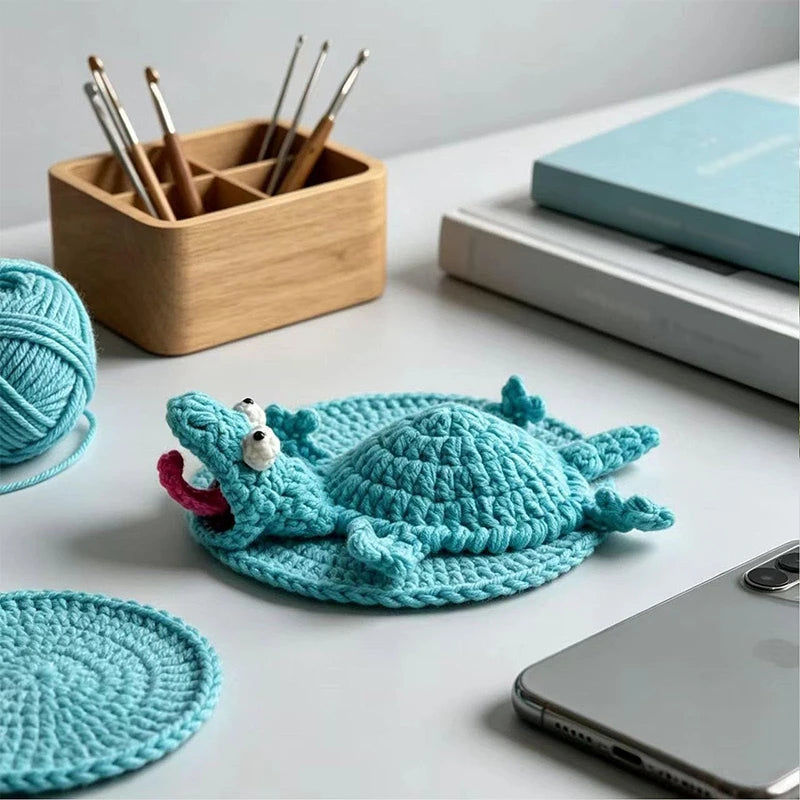 Handmade teal crochet lizard coaster resting on a white surface next to wooden tools and a smartphone.