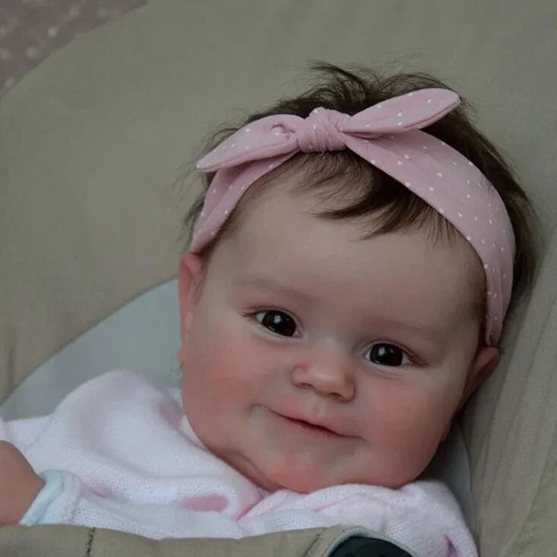 A close-up photo of a lifelike reborn baby doll with dark hair tied in a pink bow, resting on a light-colored surface.