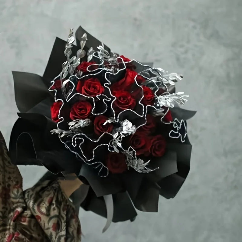 A bouquet of deep red roses wrapped in black paper with silver foliage and white string details.
