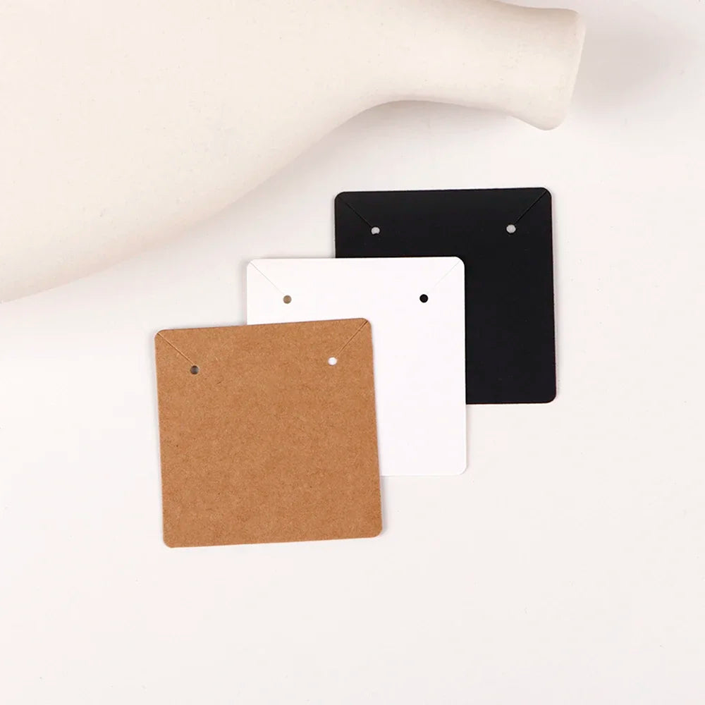 Three square jewelry display cards in brown kraft, white, and black are shown on a white surface.