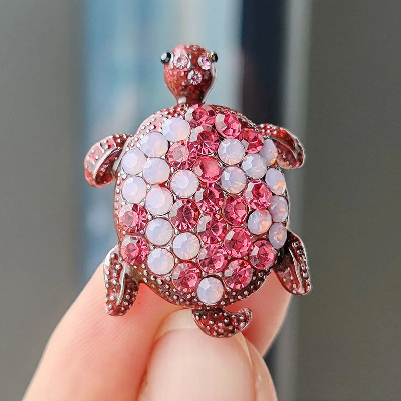 A detailed rhinestone turtle brooch pin with pink and clear stones is held in a hand.