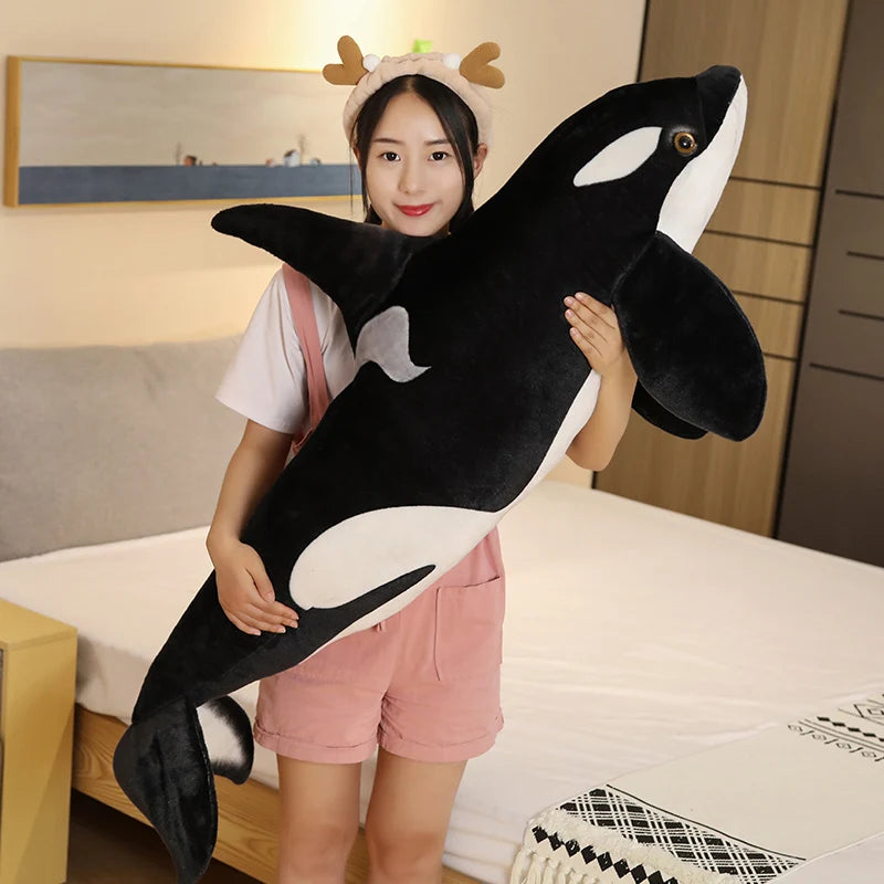A young woman is holding a large black and white orca whale plush toy in a bedroom setting.