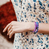 A hand wearing a stack of purple glass beads for jewelry making against a floral patterned fabric.
