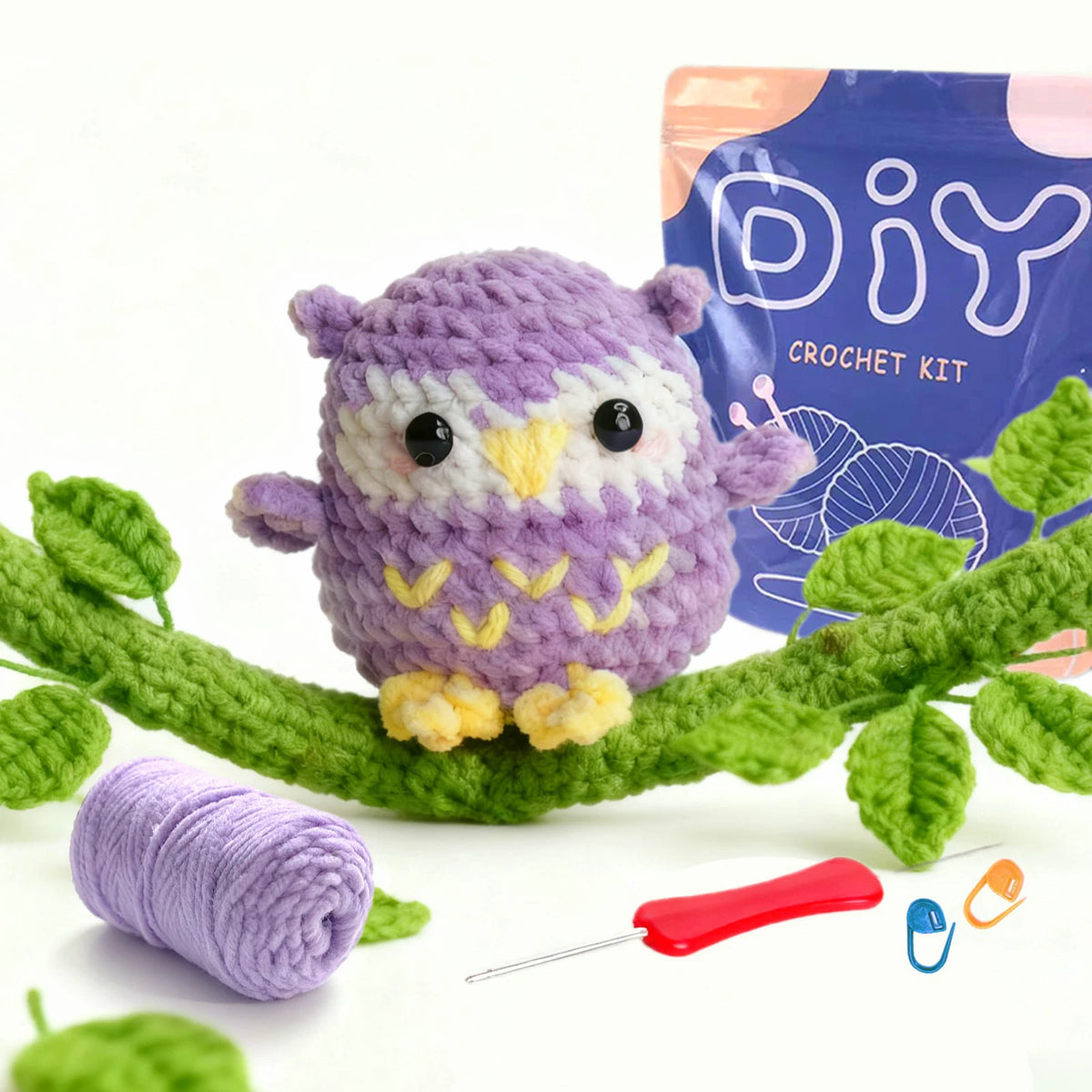 A purple crocheted owl toy sits on a green branch next to yarn and a crochet kit box.