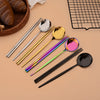 A set of six colorful stainless steel spoons and chopsticks arranged on a beige surface.