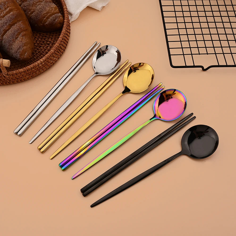 A set of six colorful stainless steel spoons and chopsticks arranged on a beige surface.