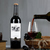 A dark glass bottle of red wine is being poured into a wine glass, with grapes and a wooden crate in the background.