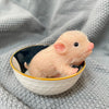 A small, pink silicone reborn piglet doll is sitting in a dark bowl on a gray knitted fabric.