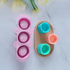 A pink silicone mold with multiple circular depressions is shown next to a wooden tray containing various colored silicone pl