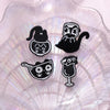 A set of four black cat enamel pins displayed on a translucent, iridescent surface.