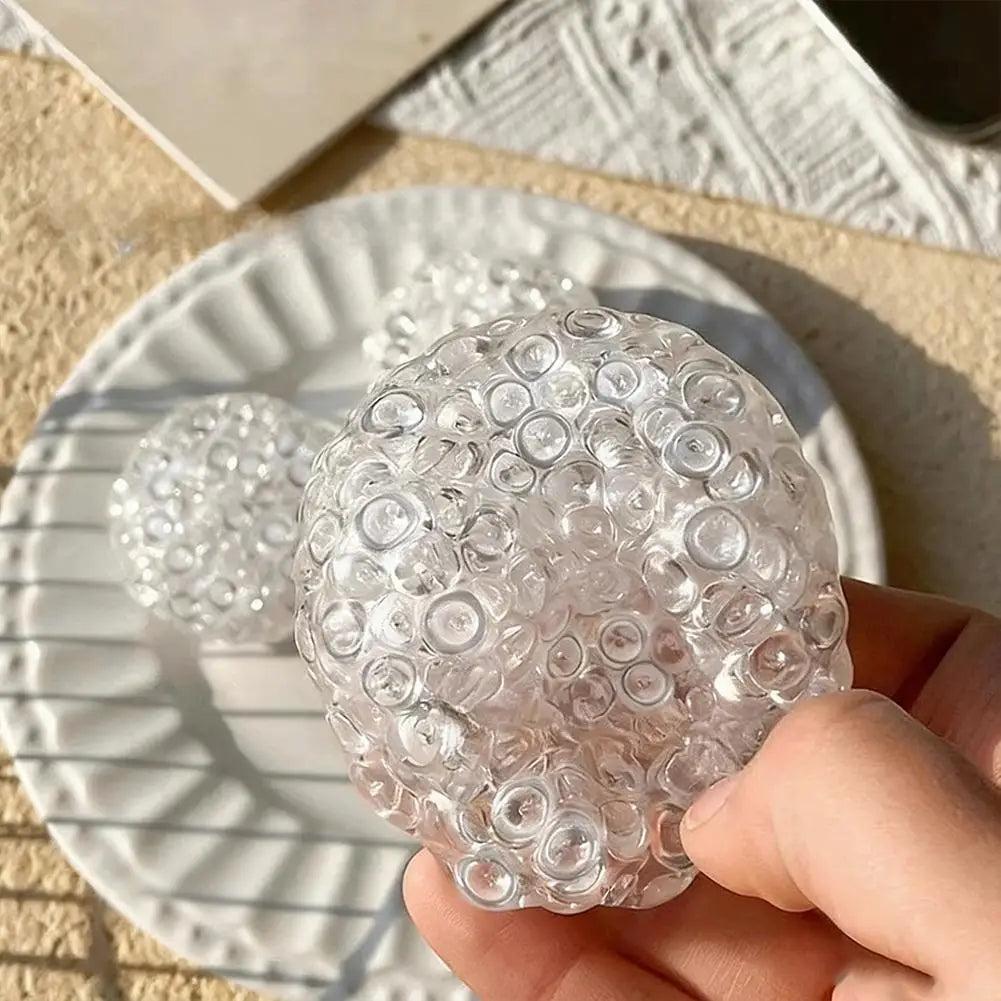 A hand holds a clear, spherical squishy bead stress ball resting on a white plate.