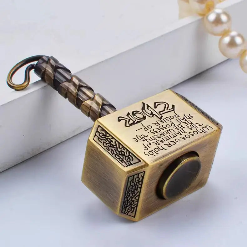 A brass hammer-shaped metal fidget spinner with a textured handle and a round top is displayed on a white surface.