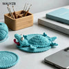 A teal handwoven crochet lizard coaster resting on a white desk next to wooden sticks and a notebook.
