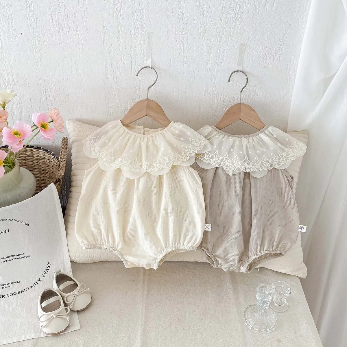 A cream-colored baby girl sleeveless jumpsuit with a lace collar hanging on a wooden hanger against a white background.