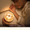 A person is gently holding a round, glowing night light projector with rotating galaxy effects in a soft indoor setting.