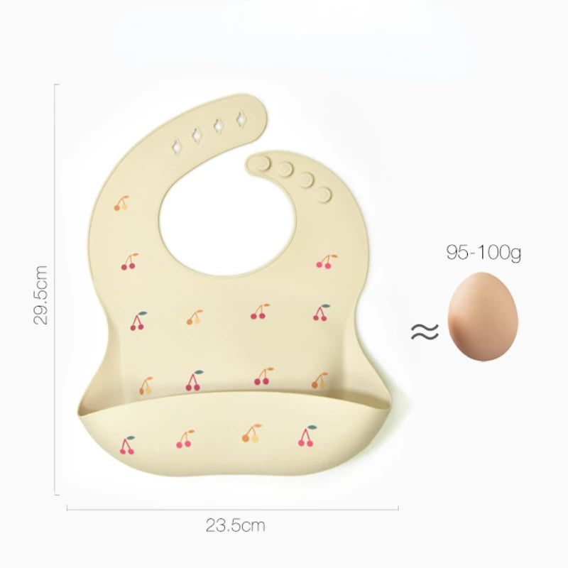 A beige silicone baby bib with an adjustable neck and small cherry pattern, shown next to an egg.