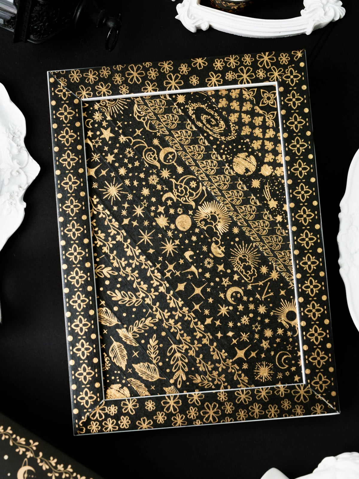 A rectangular frame displaying a dark background with intricate gold foil celestial and botanical patterns.