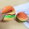 A hand holds an orange squishy hamburger stress ball with green and brown layers on a light surface.