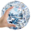 A hand places a clear watercolor sticker depicting a snowy mountain landscape and forest scene onto a blue patterned surface.