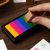 A hand holds a wooden block over a black ink pad featuring six rectangular rainbow colored pads for stamping and crafts.