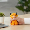 A hand is holding an orange glitter highland cow squishy stress relief toy on a wooden table.