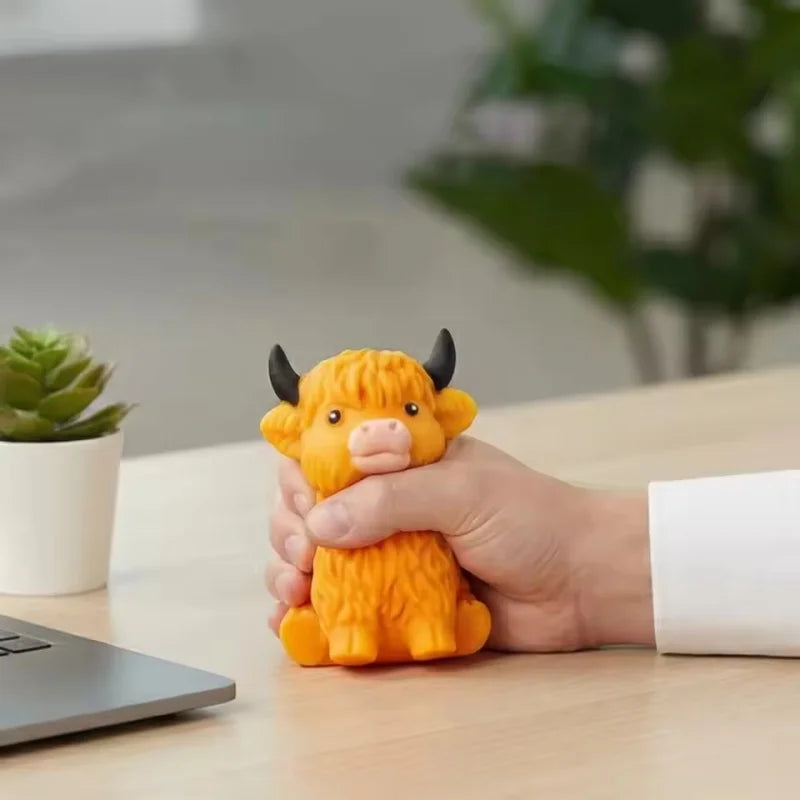 A hand is holding an orange glitter highland cow squishy stress relief toy on a wooden table.