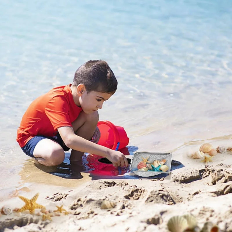 A young boy is kneeling in shallow water on a sandy beach, using a metal scoop to find treasures.