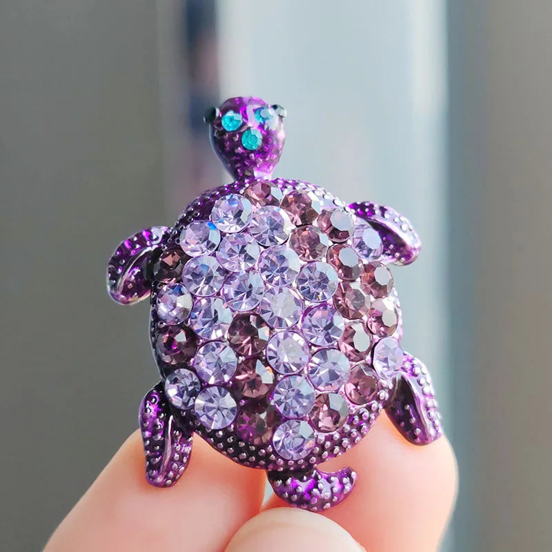 A purple rhinestone turtle brooch pin held between fingers.