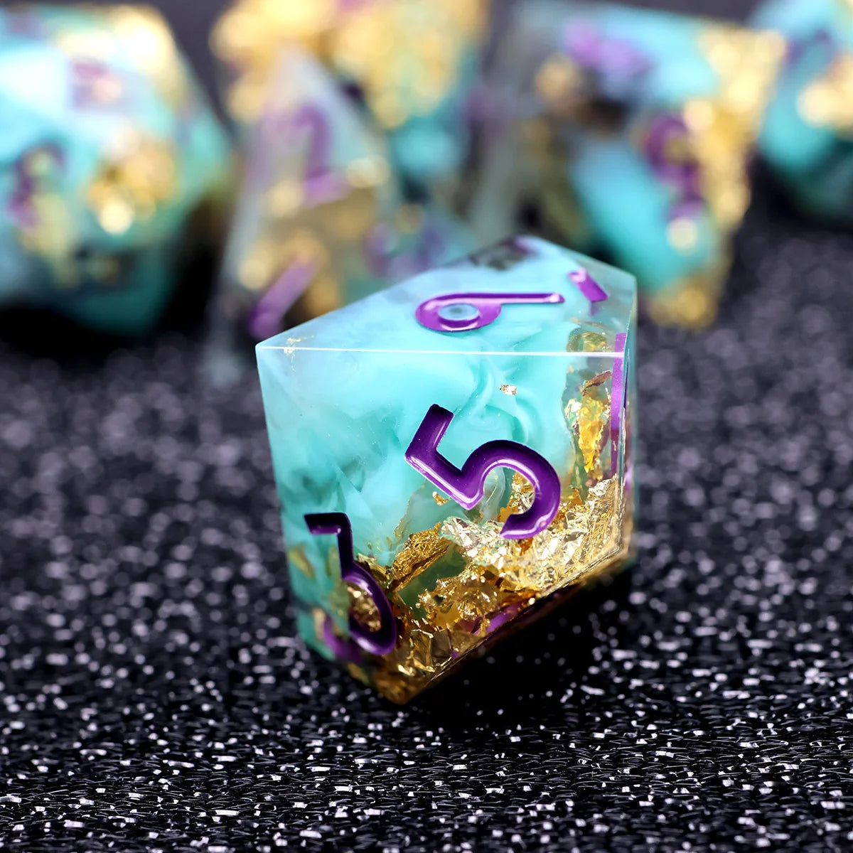 A close-up of a translucent teal resin die with purple numbers and gold glitter, resting on a dark surface.